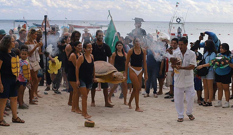 Crowd on beach with women holding turtle, man performing ritual with smoke in foreground.