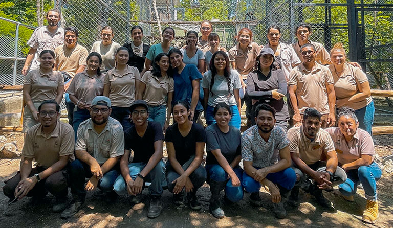 A diverse group of 25 people posing outdoors near a fence, all smiling.