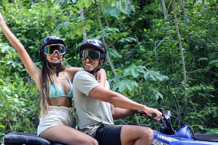 Two people on an ATV in a forest, wearing helmets and goggles, one raising an arm.