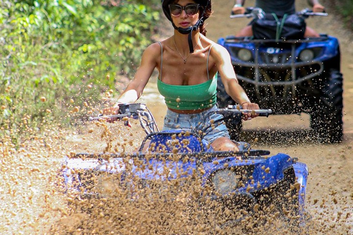 Two people on ATVs riding through mud on a forest trail.