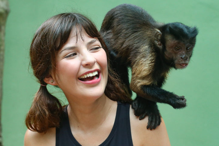 Woman with brown hair laughing as a small monkey sits on her shoulder.
