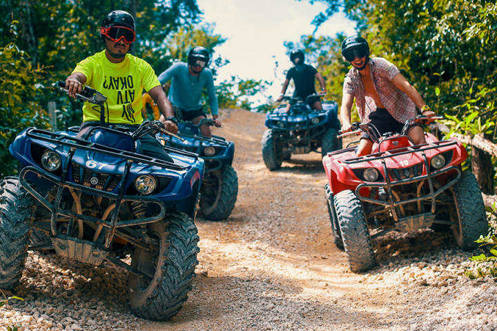 Four people riding ATVs on a dirt trail surrounded by lush greenery.