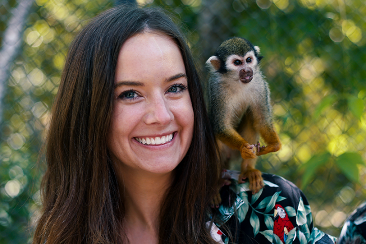 Smiling woman with long hair, monkey perched on her shoulder in a leafy area.
