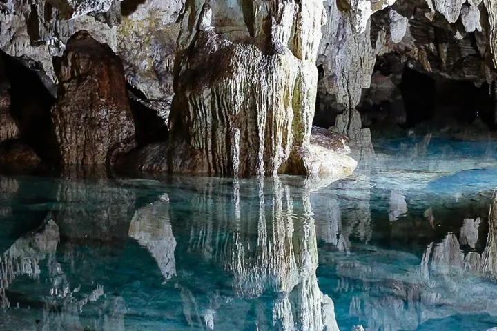 Stalactites and stalagmites in a cave with clear blue water.