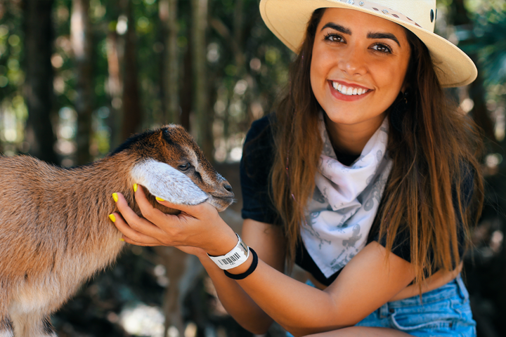 Woman in hat smiling, petting a goat in a forest setting.