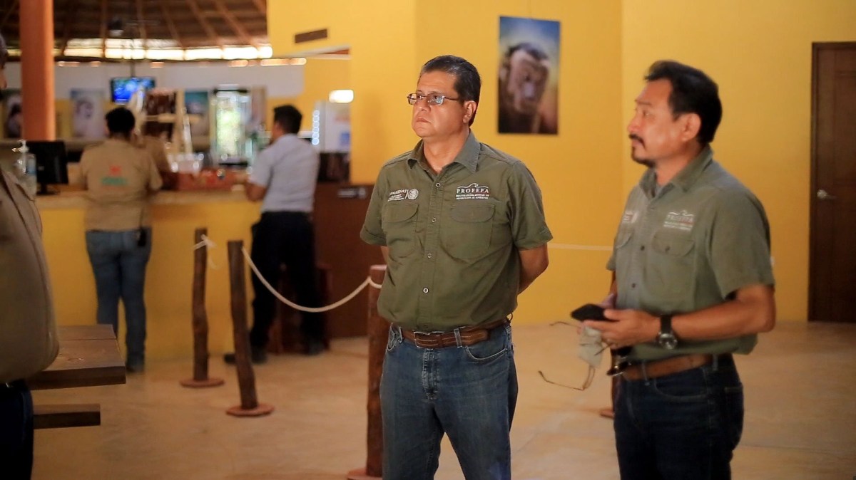 Two men in uniform shirts stand indoors with casual expressions, others busy in background.