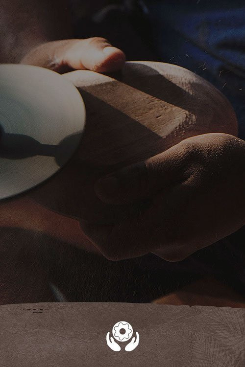 Person sanding a wooden bowl using a power tool under dim lighting.
