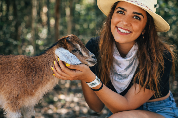 Smiling person in a hat holds a goat outdoors.