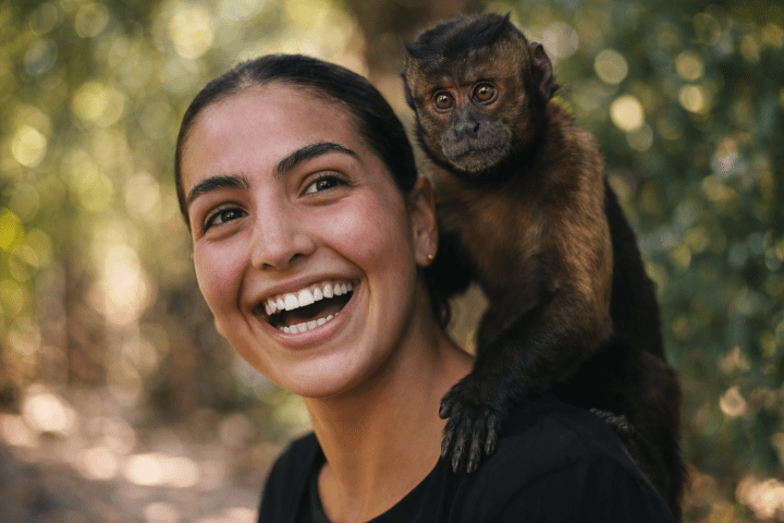 Woman smiling with a capuchin monkey on her shoulder in a forested area.