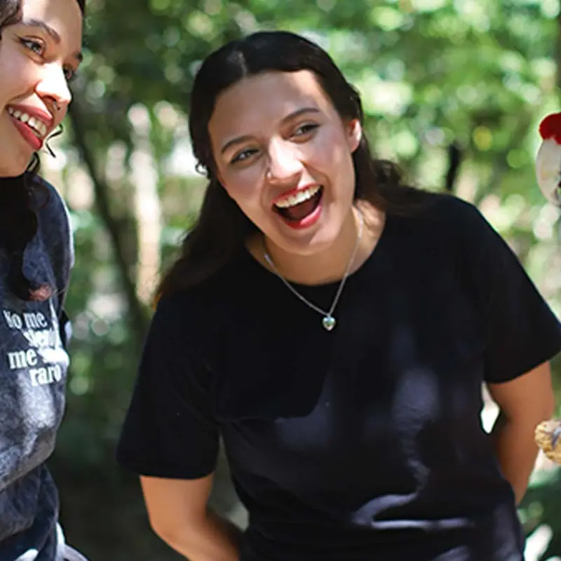 Two women smiling at a colorful parrot perched on a branch outdoors.